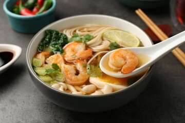 Eating delicious ramen with shrimps from bowl with spoon at grey table, closeup. Noodle soup