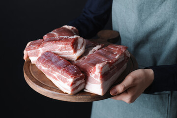 Woman holding wooden board with pieces of raw pork belly on black background, closeup