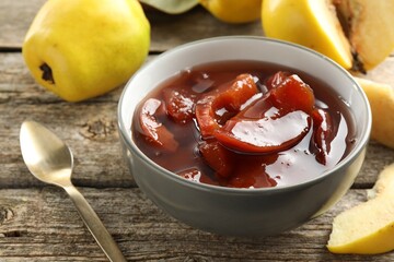 Tasty homemade quince jam in bowl, spoon and fruits on wooden table, closeup