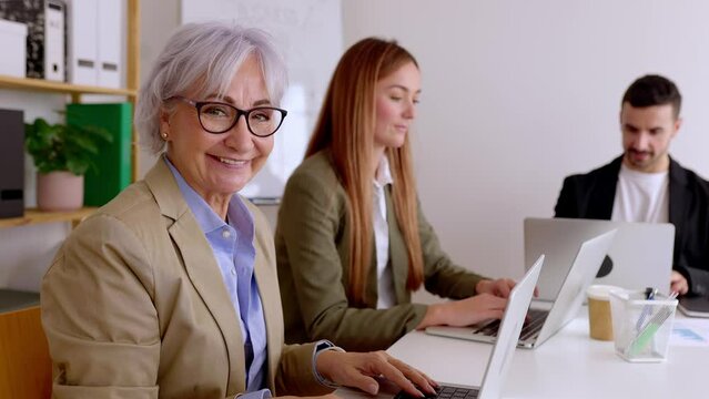 Senior businesswoman smiling at camera sitting with colleagues in the office. Joyful portrait of professional female manager working on laptop. 