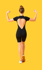 Young woman in cycling jumpsuit showing muscles on yellow background, back view