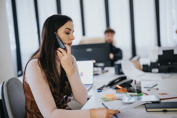 Focused young woman talking on the phone in a busy office setting, taking notes, with a coworker in the background.