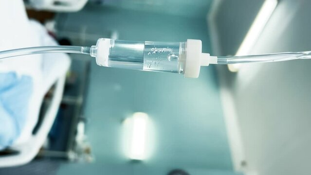 Drip chamber in an IV hanging on a medical infusion stand against the background of blurred medical beds in a clinic ward. Intravenous injections, medical care in a hospital. Vertical video