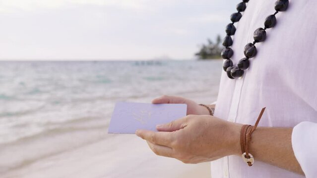 Closeup shot male hands holding embossed vow book with vow. Unrecognizable groom in boho style outfit in traditional kukui nut necklace opens vow book to read vows to bride at beach wedding ceremony