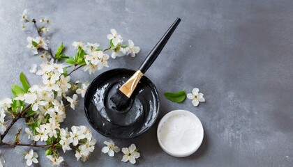 Preparing black clay cosmetic mask with almond spring flower decoration on gray background with copy space
