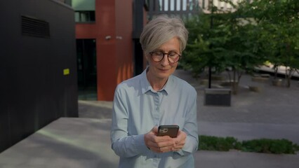 Smiling business woman elderly Caucasian mature businesswoman walking in city downtown outside texting sms messages smartphone device happy retired old female walk on street with mobile phone outdoors