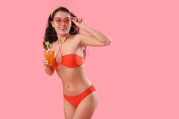 Sexy young woman in swimsuit drinking cocktail on pink background