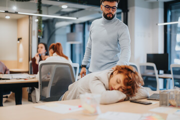 A tired employee sleeping at their working desk in the office, while a businesswoman wakes her up.