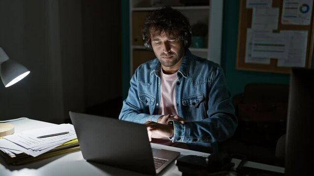 Hispanic man working late in office, wearing headphones and denim jacket, checking time, illuminated by desk lamp.