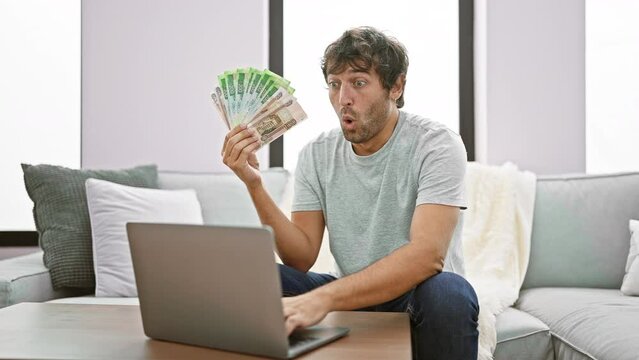 Shocked young man at home, face displaying disbelief, mouth open in amazement, handling russian rubles on laptop, expressing fear and surprise!