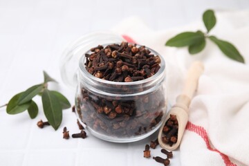 Aromatic cloves in glass jar, scoop and green leaves on white table, closeup