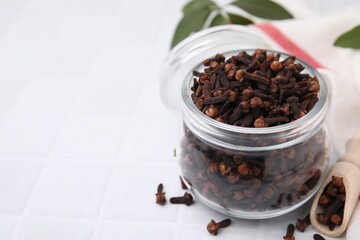 Aromatic cloves in glass jar and scoop on white tiled table, closeup. Space for text