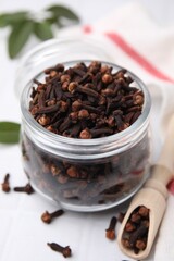 Aromatic cloves in glass jar and scoop on white table, closeup