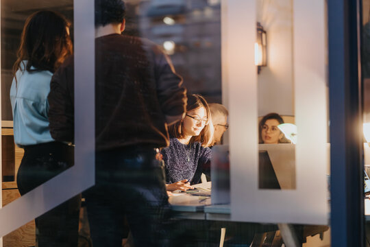 A candid shot of a diverse group of colleagues collaborating on a project during late office hours, as seen through a window. - Powered by Adobe