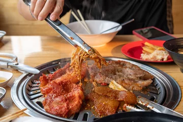 Fotobehang A woman's hand picks golden needle mushrooms marinated in sauce on a Korean wok on a grill in a restaurant. © Jonh_Walker