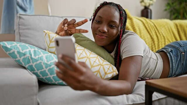 Beautiful african american woman lying on the sofa at home, joyfully taking a victory 'v' selfie with her smartphone