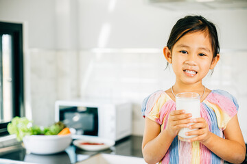 Portrait of cheerful Asian kid holding milk cup in kitchen. Smiling daughter sips enjoying the drink. Happy little girl relishing calcium-rich liquid radiating joy and happiness at home give me.