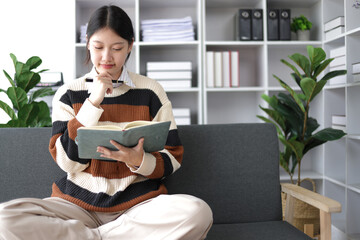 Young beautiful Asian woman reading and writing notes while sitting on sofa in living room at home.