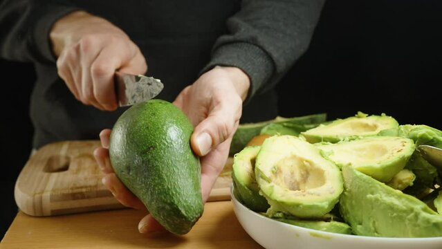 A man cuts an avocado in half, peels it, and arranges it on a plate. Against a black background. Close-up.