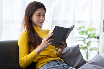 Fototapeta premium Charming Asian woman is happily reading a book on the sofa in the living room at home.