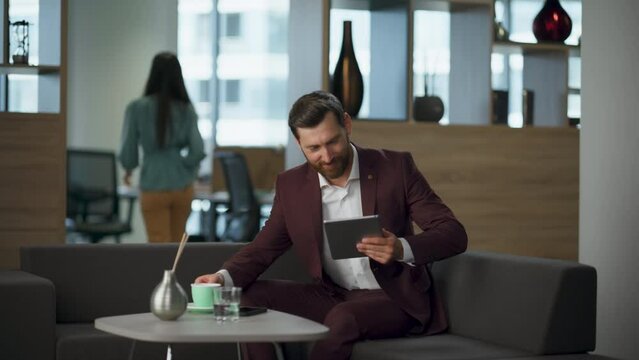 Smiling millionaire drinking coffee typing tablet in office. Relax break time
