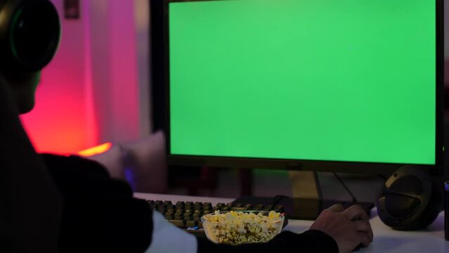 A young gamer watching the green screen - computer monitor with green chroma  watching movie. Over-the-shoulder shot of a man with chroma screen while eating popcorn - young man wearing headset  pl...
