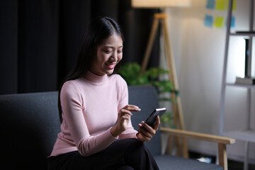 Young Asian woman sitting on sofa at home using mobile phone to send text messages and talk on the phone with her friends.