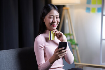 Woman sitting on sofa in living room using smartphone and credit card shopping online from home.