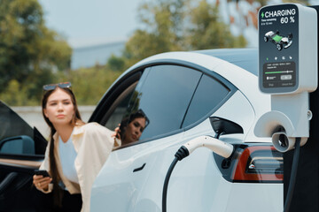 Young woman use smartphone to pay for electricity at public EV car charging station green city...