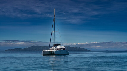 The white yacht is anchored in the ocean. A tall mast against a background of blue sky and clouds....