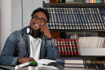 Modern African student prepares for classes in an office with books, dreamily looking up. Young...