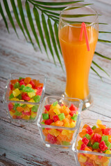 Bright sweet candied fruit is laid out in glass bowls on table, in tall glass freshly squeezed orange juice. Full glass delicious freshly squeezed juice is on table along with candied fruits in dish.