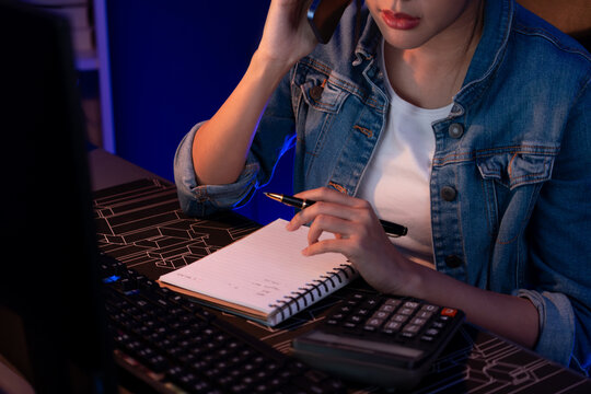 Cropped image of young Asian creative woman calling on phone to customer or coworker for marketing creator plan on pc with notebook and pen to write memo at neon modern office at night. Stratagem.