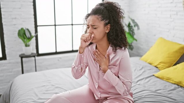 Young, beautiful hispanic woman with curly hair, seriously infected with covid-19, sits sick on her bedroom bed, coughing amidst a cold morning at home