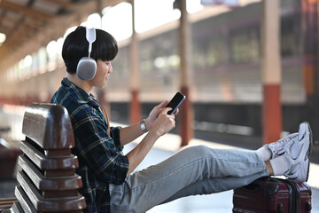 Young man in headphones using mobile phone while sitting on bench at railway station - obrazy, fototapety, plakaty