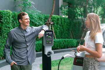 Young couple travel with EV electric car charging in green sustainable city outdoor garden in...
