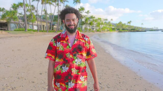 An exuberant black man with curly hair, wearing a bright Hawaiian shirt, walking backwards and waves at the camera with a friendly smile at tropical beachfront. Slow Motion, Camera 4K RAW. 