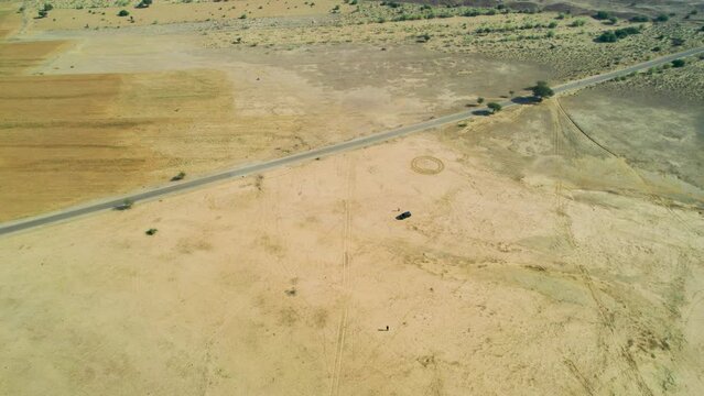 aerial drone shot orbiting car in the middle of desert with barren arid land in distance and narrow village road passing my in sam jaisalmer rajasthan India popular tourist and camping site