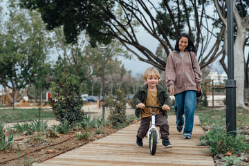 European young mother on a walk with her son on a balance bike, motherhood and joint walks around the park, Young funny mother and stylish child
