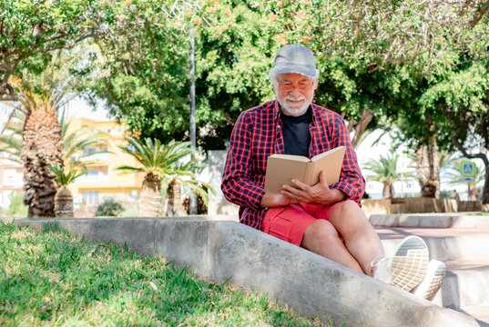 Serene retirement lifestyle. Smiling senior man sitting outdoors in public park reading a book. Bearded white-haired male with checkered shirt