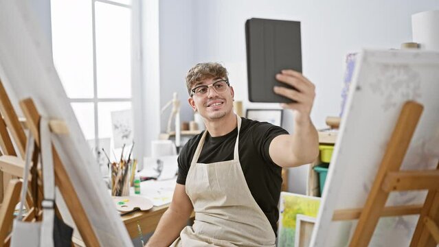 Charming young hispanic man, a budding artist, radiating joy while having a lively video call on his touchpad amidst the creative chaos of his art studio.