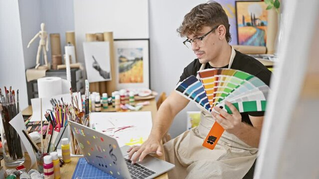 Handsome young hispanic man artist, smiling, sitting at table in art studio, choosing paint color on laptop for canvas painting