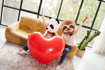 Couple of unrecognizable girls with stuffed animal masks holding giant red inflatable heart sitting...