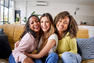 Portrait of three attractive female room mates looking smiling at camera. Group of cute cheerful girls hugging sitting on couch at home. Happy friends posing for photo indoors. Generation z and rent. - obrazy, fototapety, plakaty