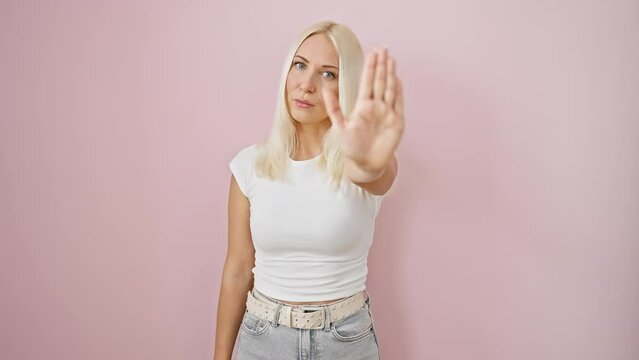 Determined young blonde woman, positively radiating 'no-go' vibes; arresting 'halt' gesture with palm; serious warning staredown on her face; standing against an isolated over-the-top pink wall.