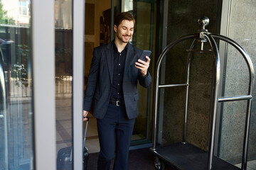 Smiling young businessman looking at smartphone while going outside hotel
