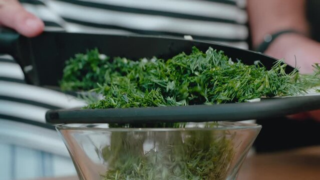 Chopped herbs and garlic being transferred from a knife to a glass bowl, with a focus on the fresh ingredients against a wooden backdrop, ideal for stock video content on cooking.