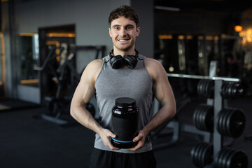 Fototapeta na wymiar Smiling man athlete posing at gym with container of protein whey, holding pack with fitness supplements, muscular guy in sport club, place for your design