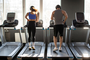 Back view of active young couple running on treadmills, man and woman performing cardio workout in...