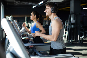 Strong man and fit woman running on treadmills or running machine in modern fitness gym, couple focusing on running, enjoying workout